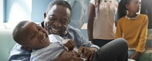 Laughing grandfather embracing grandson on a sofa while family looks on